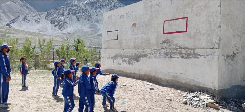 Students in blue tracksuits throwing a ball at rectangles painted on a concrete wall, mountains rising behind the school ground.