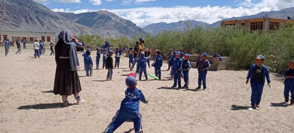 Lamdon students in blue tracksuits playing with a frisbee on an open school ground with mountains and school buildings behind.