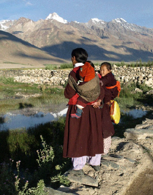 A Zanskari mother in traditional maroon dress carries a child on her back while another child walks beside her on a stone path, snow peaks behind.